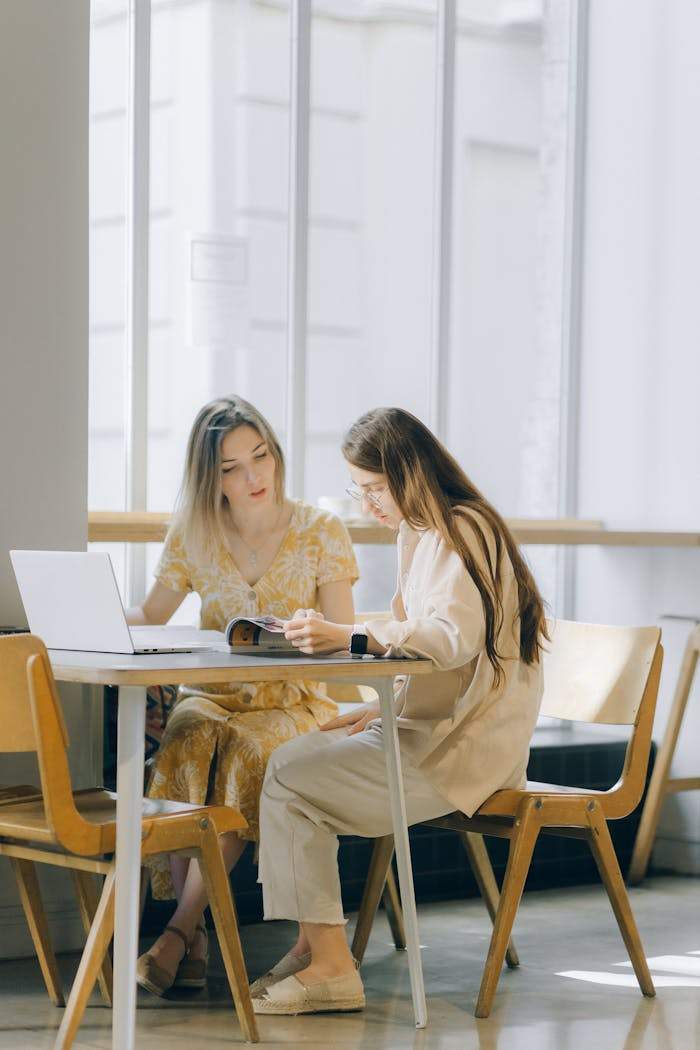 services-04 Two women collaborating over a laptop and book in a well-lit indoor setting.